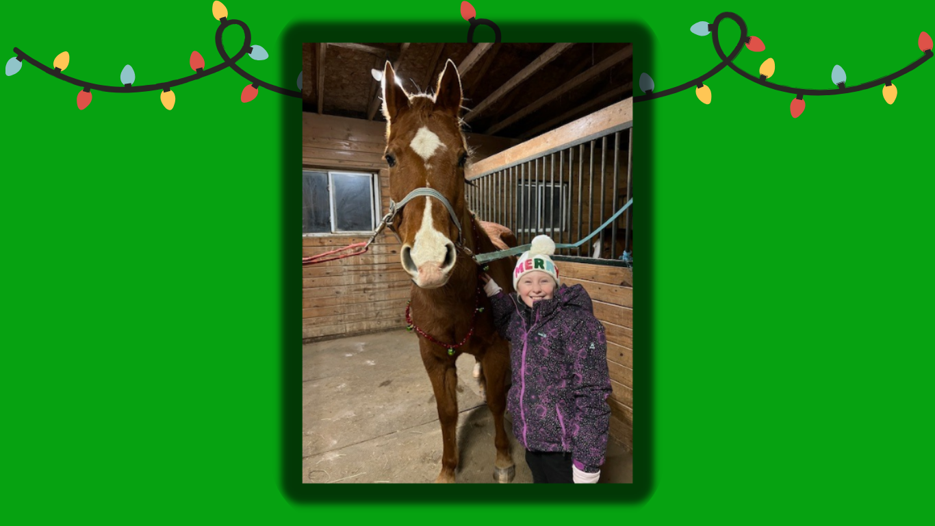 Smiling 9-year-old girl in a speckled purple and black coat wearing a white hat with the word "MERRY" in multicolor lettering poses to the right of a chestnut horse with a star and a wide snip against an indoor background of wood planks.
