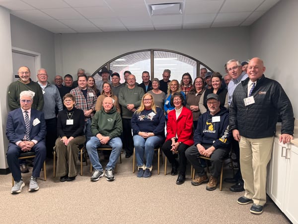 Three rows of people, some seated, pose for a picture in front of an arced window.